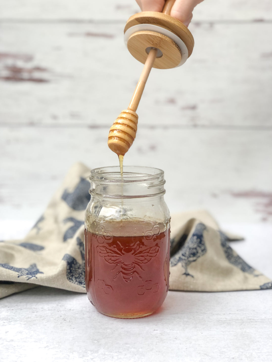 wooden honey dipper lid streaming honey into a regular mouth mason jar with chicken towel in the background